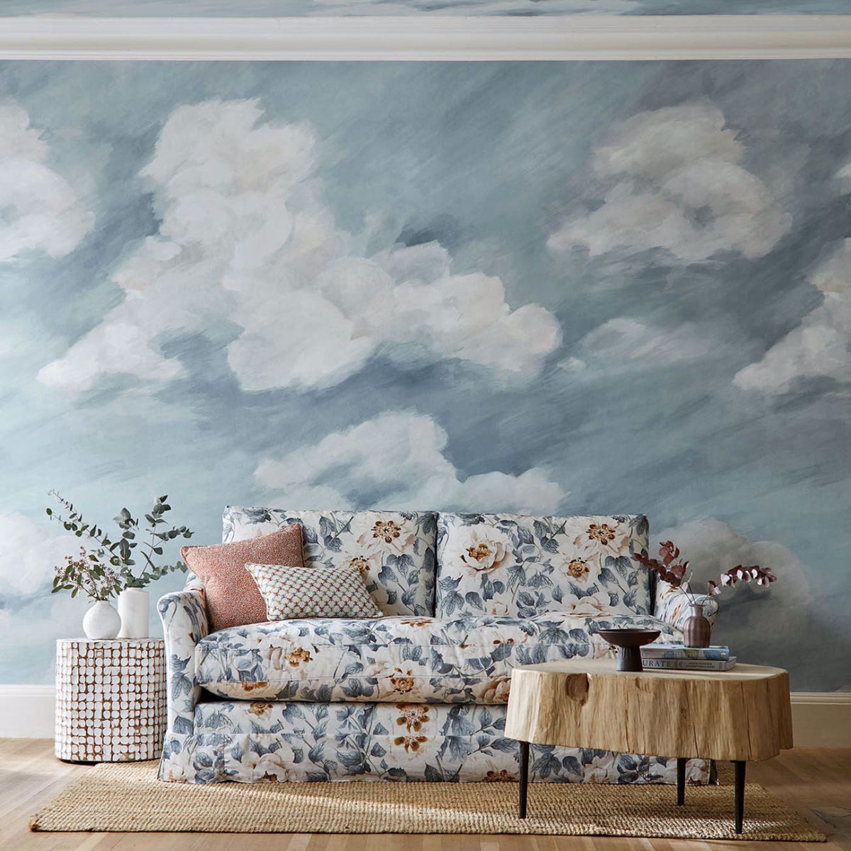 Cozy lounge or reading nook floral upholstered loveseat in front of a sky-blue wall painted with clouds, paired with a natural wood coffee table