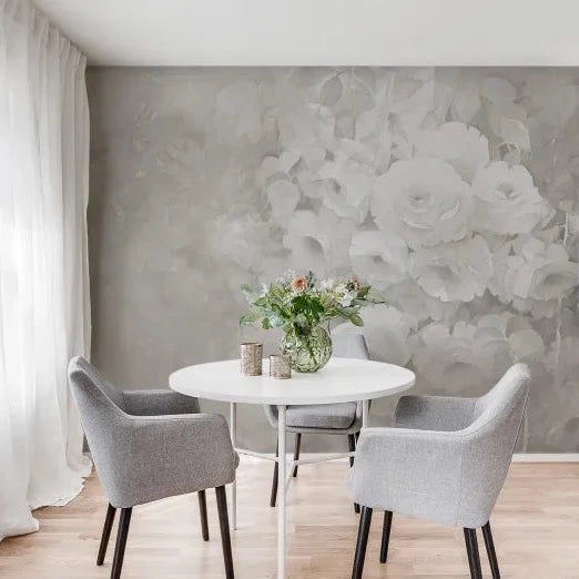 Round white table with three grey upholstered chairs, a green plant in a glass vase on top, in front of a wall with soft grey-tone wallpaper featuring a subtle rose-pattern