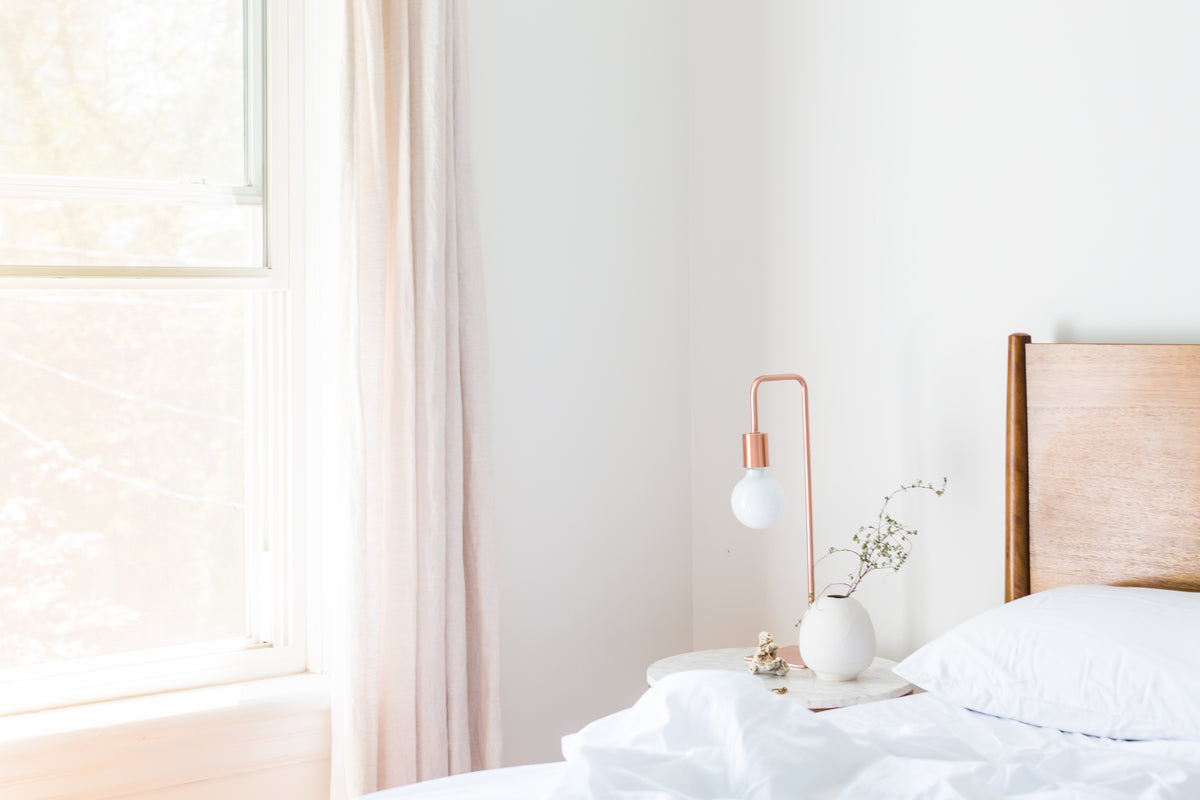 Bright bedroom corner with a copper bedside lamp on a small marble table, beside a wooden bed and white curtains softly lit by sunlight